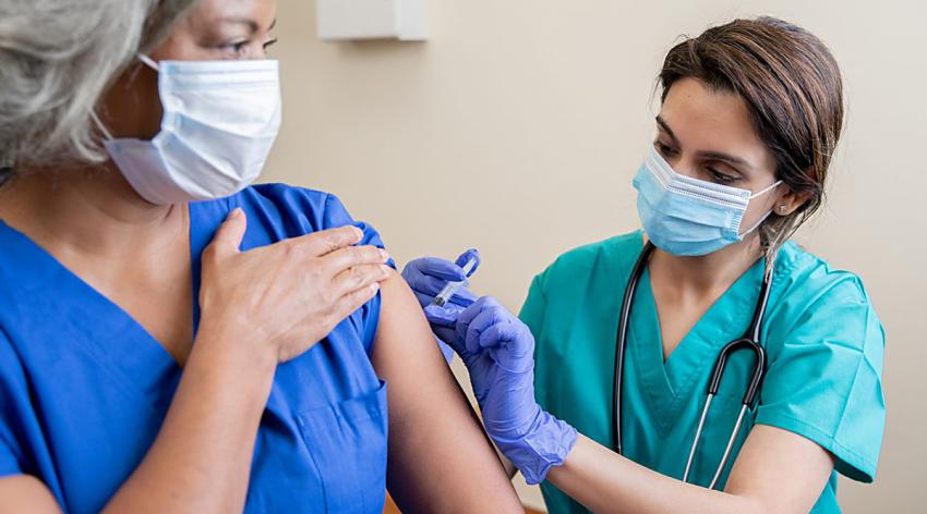 A female healthcare worker vaccinating another female healthcare worker. Both are wearing masks.